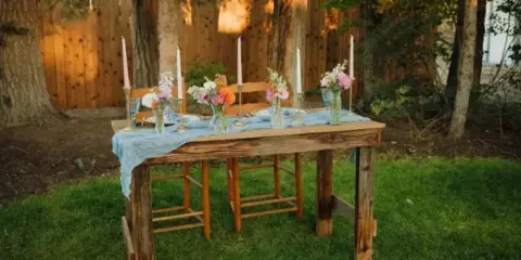 A rustic wooden table set outdoors with a blue linen runner, floral arrangements, and tall candles.