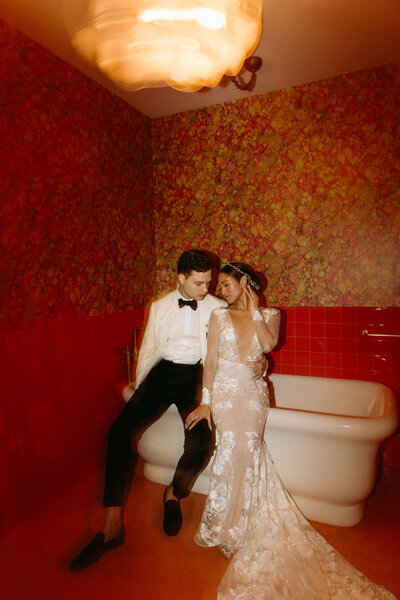 A bride and groom posing together in a room with red floral wallpaper and a white bathtub.
