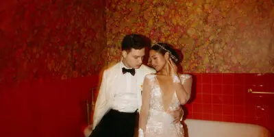 A bride and groom posing together in a room with red floral wallpaper and a white bathtub.