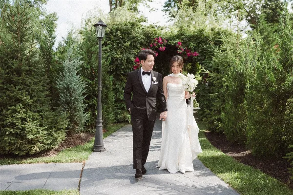A bride and groom walking hand-in-hand on a paved path through a garden with green hedges and a lamp post.