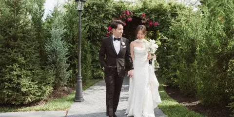 A bride and groom walking hand-in-hand on a paved path through a garden with green hedges and a lamp post.