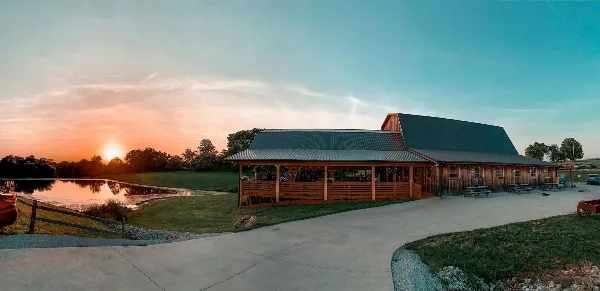 Exterior view of a rustic wooden barn wedding venue with a covered patio and a pond at sunset.