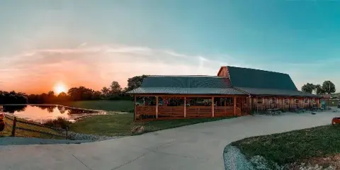 Exterior view of a rustic wooden barn wedding venue with a covered patio and a pond at sunset.