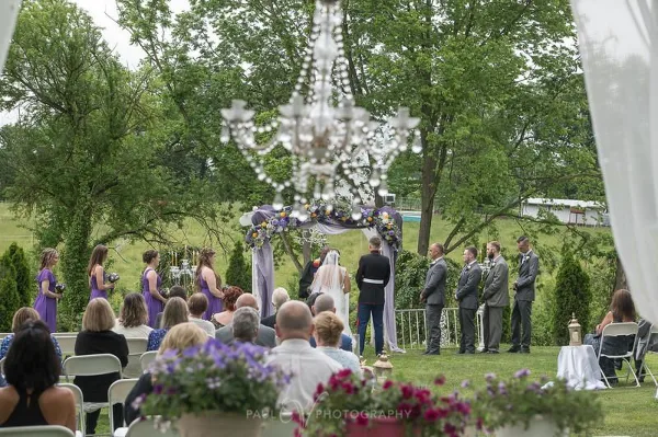 Outdoor garden wedding ceremony with a floral arch, hanging chandelier, seated guests, and a couple at the altar.
