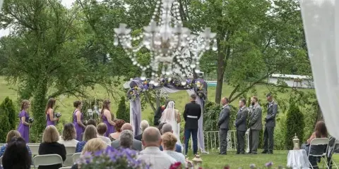 Outdoor garden wedding ceremony with a floral arch, hanging chandelier, seated guests, and a couple at the altar.