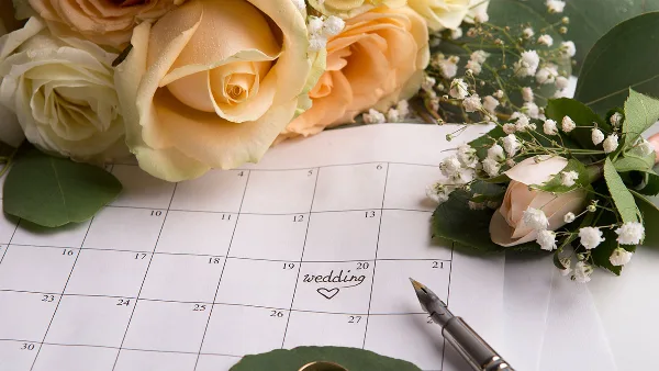 A close-up of a calendar with the word 'wedding' written on a date, surrounded by a bouquet of cream roses and baby's breath, with a fountain pen resting on the page.