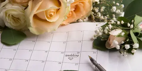 A close-up of a calendar with the word 'wedding' written on a date, surrounded by a bouquet of cream roses and baby's breath, with a fountain pen resting on the page.