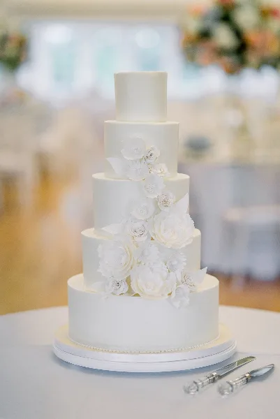 A five-tier white wedding cake with cascading white sugar flowers on a table in a banquet hall.