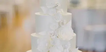 A five-tier white wedding cake with cascading white sugar flowers on a table in a banquet hall.