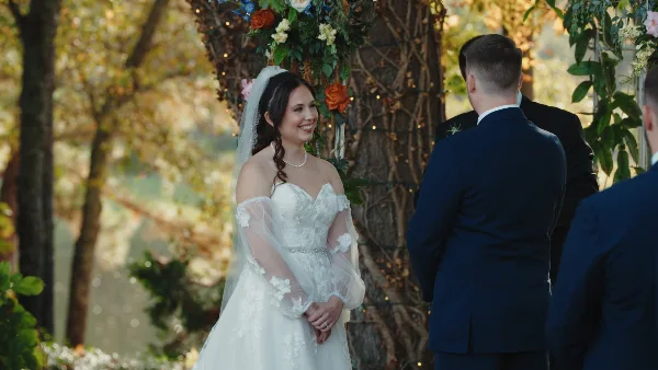 A bride in a white lace gown and veil smiles during an outdoor wedding ceremony under a floral arch, with the groom in a navy suit facing her.