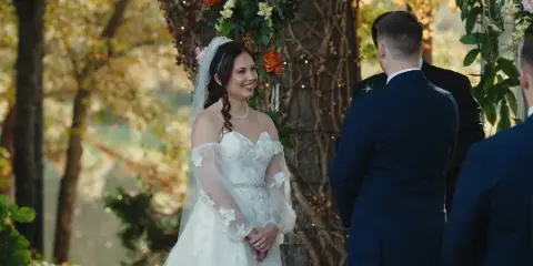 A bride in a white lace gown and veil smiles during an outdoor wedding ceremony under a floral arch, with the groom in a navy suit facing her.