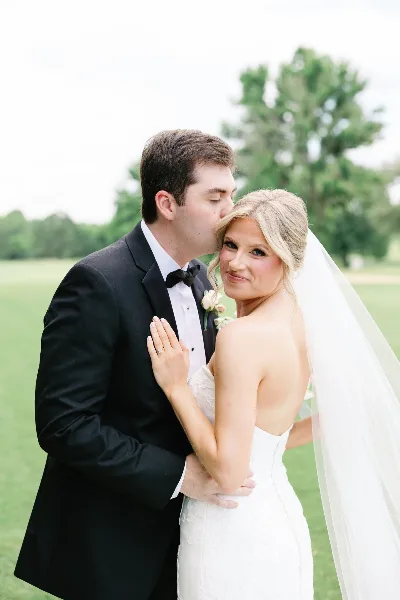 A bride and groom sharing a kiss in a field, with the groom in a black tuxedo and the bride in a white wedding dress.