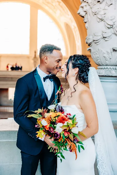 A bride and groom in formal wedding attire share a close moment while holding a vibrant bouquet in an elegant architectural setting.
