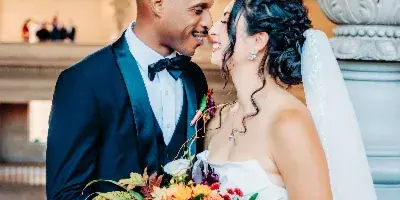A bride and groom in formal wedding attire share a close moment while holding a vibrant bouquet in an elegant architectural setting.