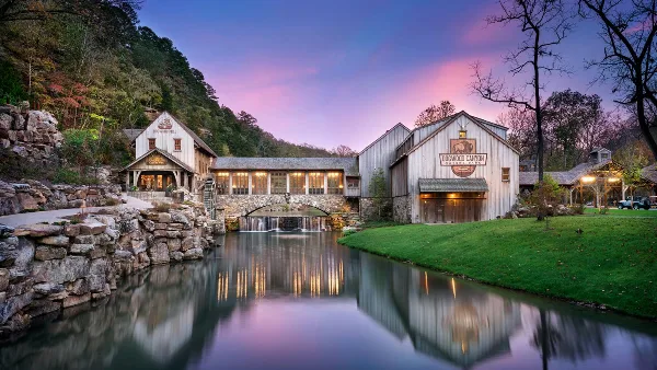 A scenic view of the Dogwood Canyon Nature Park entrance buildings, featuring a stone bridge over a creek and a pink sunset sky.