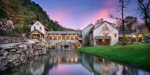 A scenic view of the Dogwood Canyon Nature Park entrance buildings, featuring a stone bridge over a creek and a pink sunset sky.