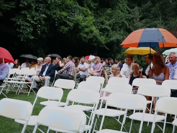 Outdoor wedding ceremony with guests seated in white chairs on a lawn, some holding colorful umbrellas.