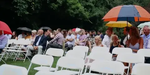 Outdoor wedding ceremony with guests seated in white chairs on a lawn, some holding colorful umbrellas.