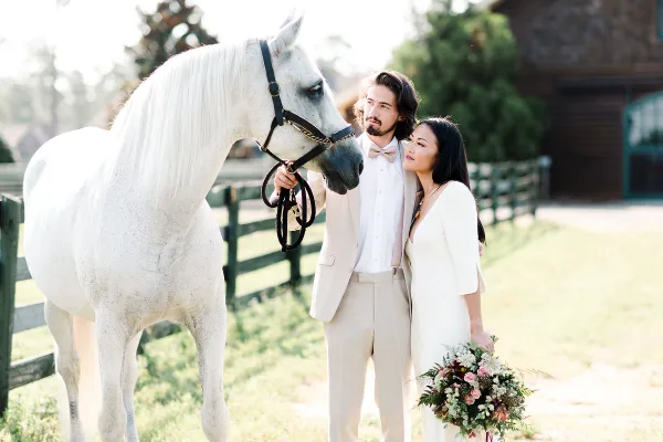 A bride and groom in formal attire posing with a white horse in a grassy outdoor setting.