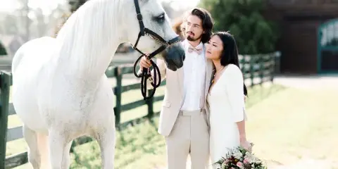 A bride and groom in formal attire posing with a white horse in a grassy outdoor setting.