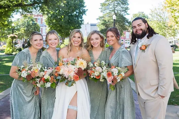 A bride in a white gown stands with four bridesmaids in green dresses and a groom in a beige suit, all holding bouquets and smiling outdoors.