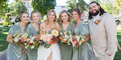 A bride in a white gown stands with four bridesmaids in green dresses and a groom in a beige suit, all holding bouquets and smiling outdoors.