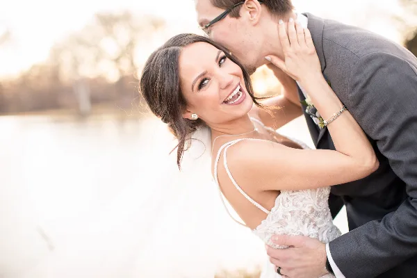 A bride in a white lace gown smiling while a groom in a grey suit kisses her cheek, set against a blurred outdoor background.