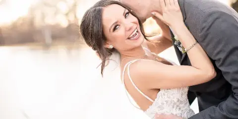 A bride in a white lace gown smiling while a groom in a grey suit kisses her cheek, set against a blurred outdoor background.