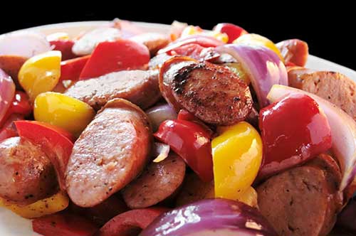A close-up photograph of a plated dish featuring sliced sausage, red and yellow bell peppers, and red onions.