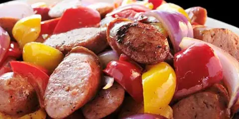 A close-up photograph of a plated dish featuring sliced sausage, red and yellow bell peppers, and red onions.