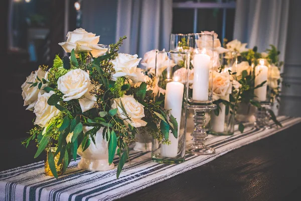 Wedding table decorated with white rose floral arrangements and lit pillar candles in glass holders.