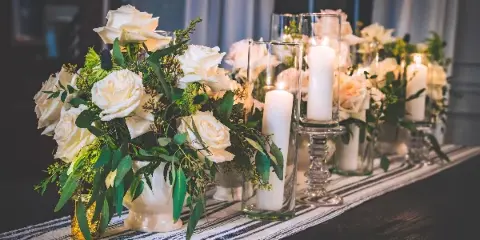 Wedding table decorated with white rose floral arrangements and lit pillar candles in glass holders.