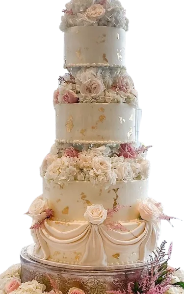 A four-tier white wedding cake decorated with fresh roses, gold leaf accents, and a draped fondant base.