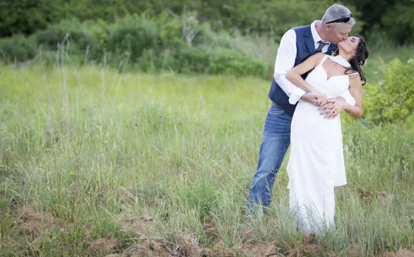 A bride in a white dress and a groom in a vest and jeans share a kiss in a grassy field.