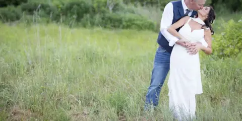 A bride in a white dress and a groom in a vest and jeans share a kiss in a grassy field.