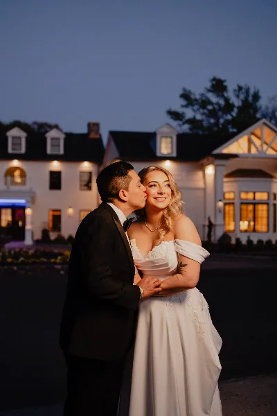 A bride and groom embracing in front of a white estate building at dusk.