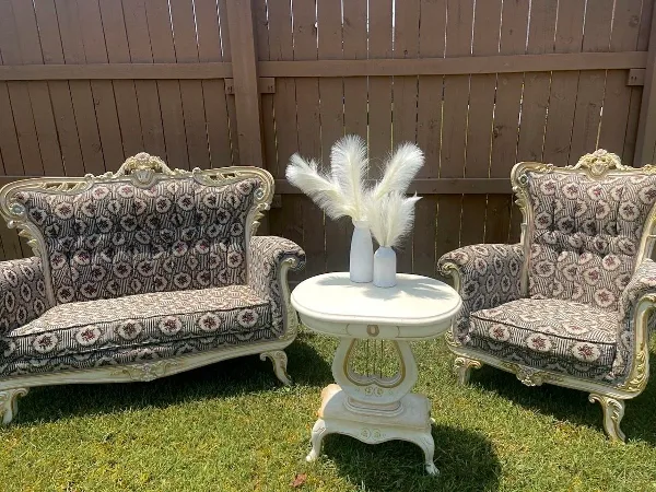 A vintage-style settee and armchair with patterned upholstery and gold accents, arranged around a white round side table with a vase of pampas grass.
