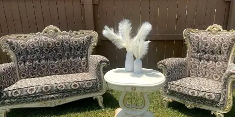 A vintage-style settee and armchair with patterned upholstery and gold accents, arranged around a white round side table with a vase of pampas grass.