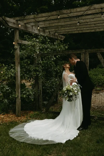 A bride and groom sharing an intimate moment under a wooden pergola with string lights and greenery.