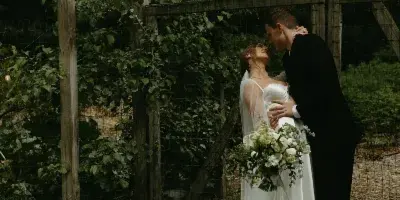 A bride and groom sharing an intimate moment under a wooden pergola with string lights and greenery.