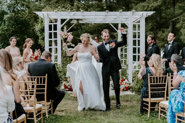 A bride and groom walking down the aisle at an outdoor wedding ceremony, surrounded by guests and floral arrangements.
