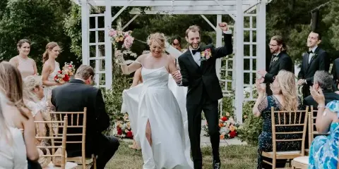 A bride and groom walking down the aisle at an outdoor wedding ceremony, surrounded by guests and floral arrangements.