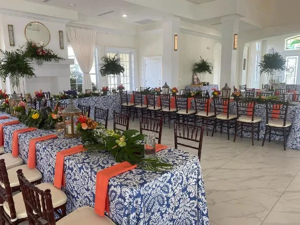 An indoor event space set up with long tables covered in blue and white patterned linens, orange sashes, floral centerpieces, and rows of dark wood Chiavari chairs.