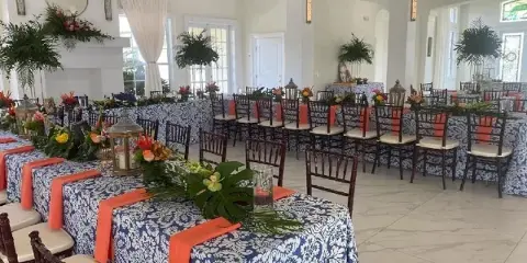 An indoor event space set up with long tables covered in blue and white patterned linens, orange sashes, floral centerpieces, and rows of dark wood Chiavari chairs.
