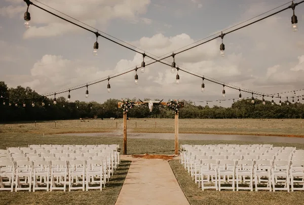Outdoor wedding ceremony setup with white chairs, a rustic wooden arch with flowers and a longhorn skull, string lights, and a rural pasture backdrop.