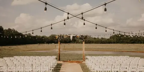 Outdoor wedding ceremony setup with white chairs, a rustic wooden arch with flowers and a longhorn skull, string lights, and a rural pasture backdrop.
