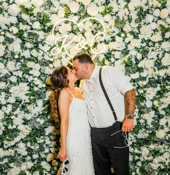 A bride and groom kissing in front of a white flower wall and a neon heart sign.