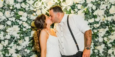 A bride and groom kissing in front of a white flower wall and a neon heart sign.