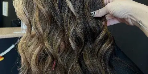 Stylist's hand adjusting a client's wavy, highlighted hair in a salon setting.