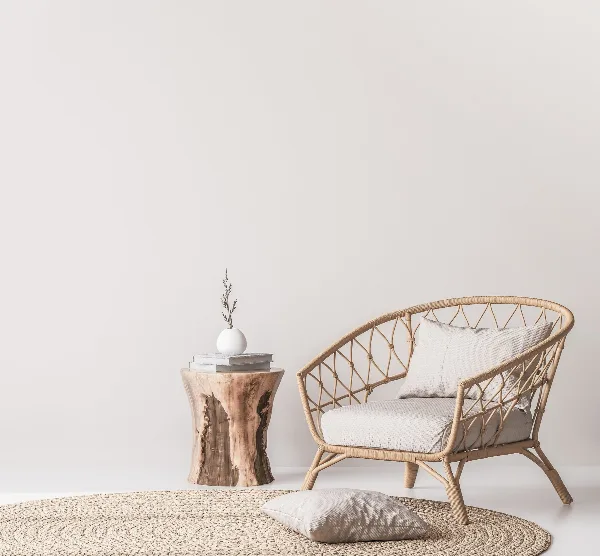 A rattan armchair with a cushion, a wooden stump side table with a vase, and a woven rug arranged in a minimalist setting.